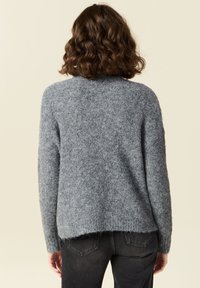 Maglione grigio lavorato a maglia con una finitura morbida e testurizzata, caratterizzato da un orlo posteriore leggermente più lungo e polsini a coste. Indossato con jeans scuri.