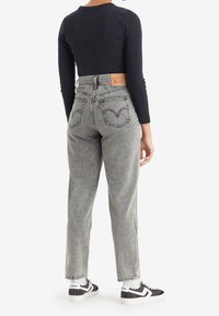 Persona con jeans grises de tiro alto de Levi's, camiseta de manga larga negra, calcetines blancos y zapatillas de Levi's en blanco y negro, de pie con la espalda hacia la cámara.