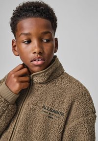 Brun fleecejakke med høj krave, struktureret stof og lynlåslukning; har "ALLSAINTS" broderet med mørk tråd.