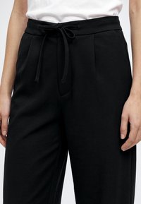 Pantalon noir taille haute avec cordon noué en nœud, porté avec un top blanc, montrant la taille jusqu'à mi-cuisse et les mains détendues sur les côtés.