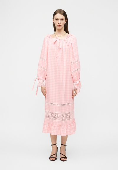 Femme debout portant une longue robe en vichy rose avec des inserts en dentelle, des manches bouffantes attachées aux poignets, et des talons noirs à brides.