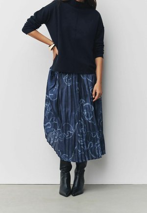 Femme portant un haut noir à manches longues, une jupe plissée bleu marine avec des motifs de cordes, des bottes noires à talons et un bracelet à son poignet gauche.
