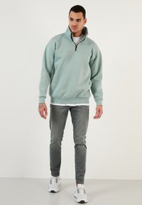 Sudadera de media cremallera en verde azulado claro con una textura suave, corte relajado, puños y dobladillo acanalados, combinada con jeans grises y zapatillas blancas.