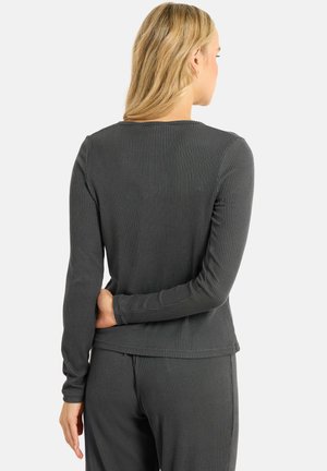 Frau mit blondem Haar, die ein dunkelgraues, geripptes Langarmshirt und passende gerippte Hose trägt, von hinten vor einem schlichten Hintergrund gezeigt.