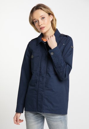 Jeune femme portant une veste bleu marine boutonnée avec des poches poitrine et un jean bleu clair, posant devant un fond blanc uni.