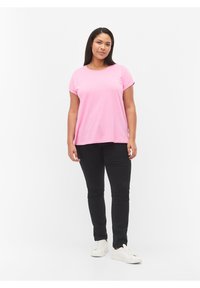 Camiseta rosa de manga corta con puños enrollados, combinada con pantalones ajustados negros y zapatillas blancas, de pie frente a un fondo neutral.