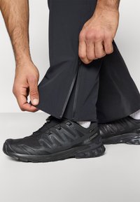 Μαύρο αθλητικό παντελόνι με φερμουάρ στον αστράγαλο. Διακρίνεται ένα μαύρο παπούτσι για ορεινό τρέξιμο με υφασμάτινη υφή και λαστιχένια σόλα.