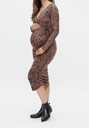 Zwangere vrouw in een bruin-zwarte patterned lange mouwen jurk die haar buik vasthoudt, met zwarte enkellaarsjes op een witte achtergrond.