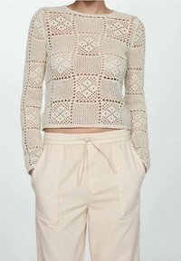 Krémový, zkrácený pletený svetr s jemným otvoreným vzorem, kulatým výstřihem a dlouhými rukávy, v kombinaci se světle béžovými kalhotami s elastickým pasem.