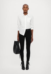 Camisa blanca de botón, jeans ajustados negros y botas de tacón negro hasta el tobillo. Sosteniendo una bolsa de mano negra. Texturas suaves, diseño ajustado.