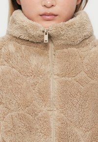Nærbillede af en person iført en beige, fluffy zip-jakke med en høj krave, der dækker nakken.