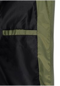 Interior de chaqueta en negro y verde oliva, con un acabado satinado suave y un bolsillo delineado en verde oliva sobre un fondo negro.