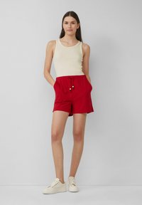 Pantaloni corti di cotone rossi con vita elastica e cordoncino, caratterizzati da dettagli in legno. Abbinati a una canottiera beige e sneakers bianche.