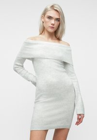 Kvinne med lyseblondt hår iført en lys grå off-shoulder strikkekjole, står med den ene hånden på hoften mot en enkel hvit bakgrunn.