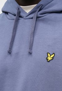 Oljčna vijolična jopa iz bombažne mešanice tkanine z žepom kengurujčka, nastavljivimi vrvicami in rumenim vezenim logotipom ptice na prsih.