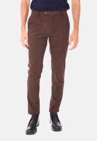 Uomo che indossa pantaloni in velluto a coste marrone e scarpe stringate in pelle nera, in piedi contro uno sfondo chiaro e neutro.