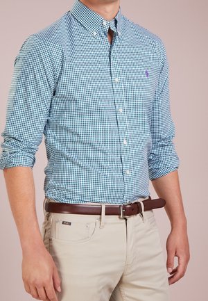 Mann trägt ein langärmeliges blau-weiß kariertes Hemd, in beige Hose gesteckt, mit braunem Ledergürtel, steht vor schlichtem Hintergrund.