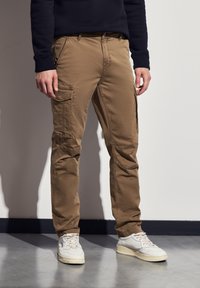 Khaki cargo kalhoty vyrobené z bavlny, s několika bočními kapsami, zúženými nohavicemi a úzkým střihem. Nosené s bílimi teniskami.