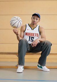 Basketbalista v bielom drese s červenými akcentmi, sedí na lavičke, drží basketbal, nosí modrú čapicu a džínsy s bielymi topánkami.