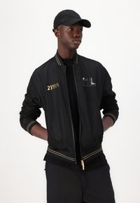 Black bomber jacket with gold accents, zip closure, and embroidered details. Paired with a black cap and black shirt beneath.