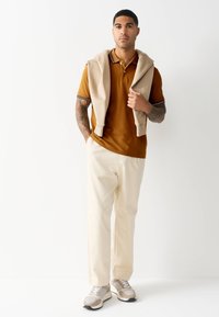 Hnedé polo tričko s krátkymi rukávmi a pásikavým golierom, béžový sveter prehodený cez ramená, biele nohavice a tenisky v neutrálnych tónoch.