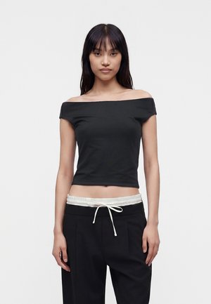 Jonge vrouw met lang zwart haar draagt een aangesloten zwarte off-shoulder crop top en zwarte broek met witte trekkoord tailleband.