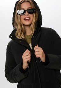 Sort hættejakke med lynlås og snore. Modellen har en strikket grøn sweater på nedenunder. Akkompagneret af sorte solbriller.