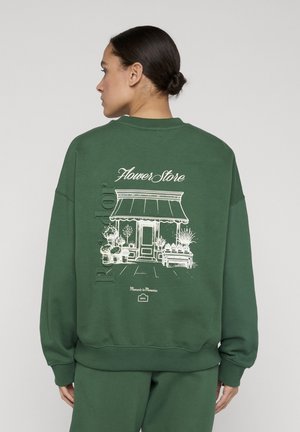 Kvinde med opsat hår iført grøn sweatshirt med hvid blomsterbutik-illustration og tekst på ryggen, stående mod en grå baggrund.