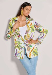 Frau in einem weißen Blazer mit bunten Blumenmustern, weißem Top, hellblauen Jeans und gelben Quastenohrringen, posiert mit geschlossenen Augen.