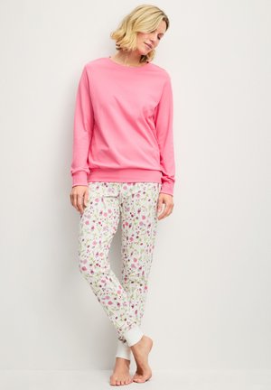 Woman standing barefoot wearing a plain pink long-sleeve top and white pants with pink and green floral print against a plain background.