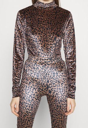 Femme portant un body à manches longues et col haut imprimé léopard avec un pantalon ajusté assorti, debout devant un fond clair uni.