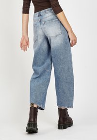 Person, der hellblaue weit geschnittene Jeans und dunkelbraune Schnürstiefel trägt, mit dem Rücken zur Kamera stehend vor einem neutralen Hintergrund.