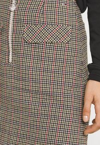 Geruite jurk met groene, zwarte en rode strepen. Bevat een ritssluiting en een zak met een diagonaal patroon. Gevoel van textuur is zichtbaar.