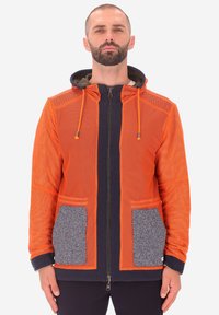 Uomo che indossa una felpa con zip arancione in rete con bordi blu navy e tasche frontali grigie texturizzate, in piedi davanti a uno sfondo bianco semplice.