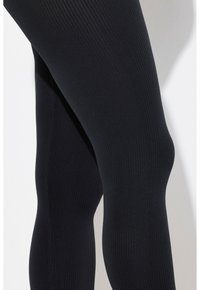 Leggings noirs à côtes, présentant un design ajusté, une texture lisse et une taille haute qui se fond parfaitement dans la silhouette des jambes.