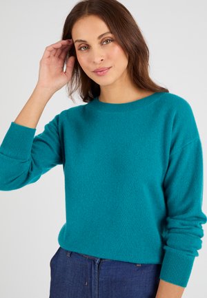 Mujer con cabello castaño que lleva un suéter de manga larga color verde azulado y vaqueros azules, levantando la mano derecha cerca de su rostro sobre un fondo liso.
