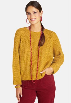 Lächelnde Frau, die einen senfgelben Strickpullover, rote Hose, rote Perlenkette, rote Creolen und goldene Armbänder trägt, mit seitlich geflochtenem Haar.