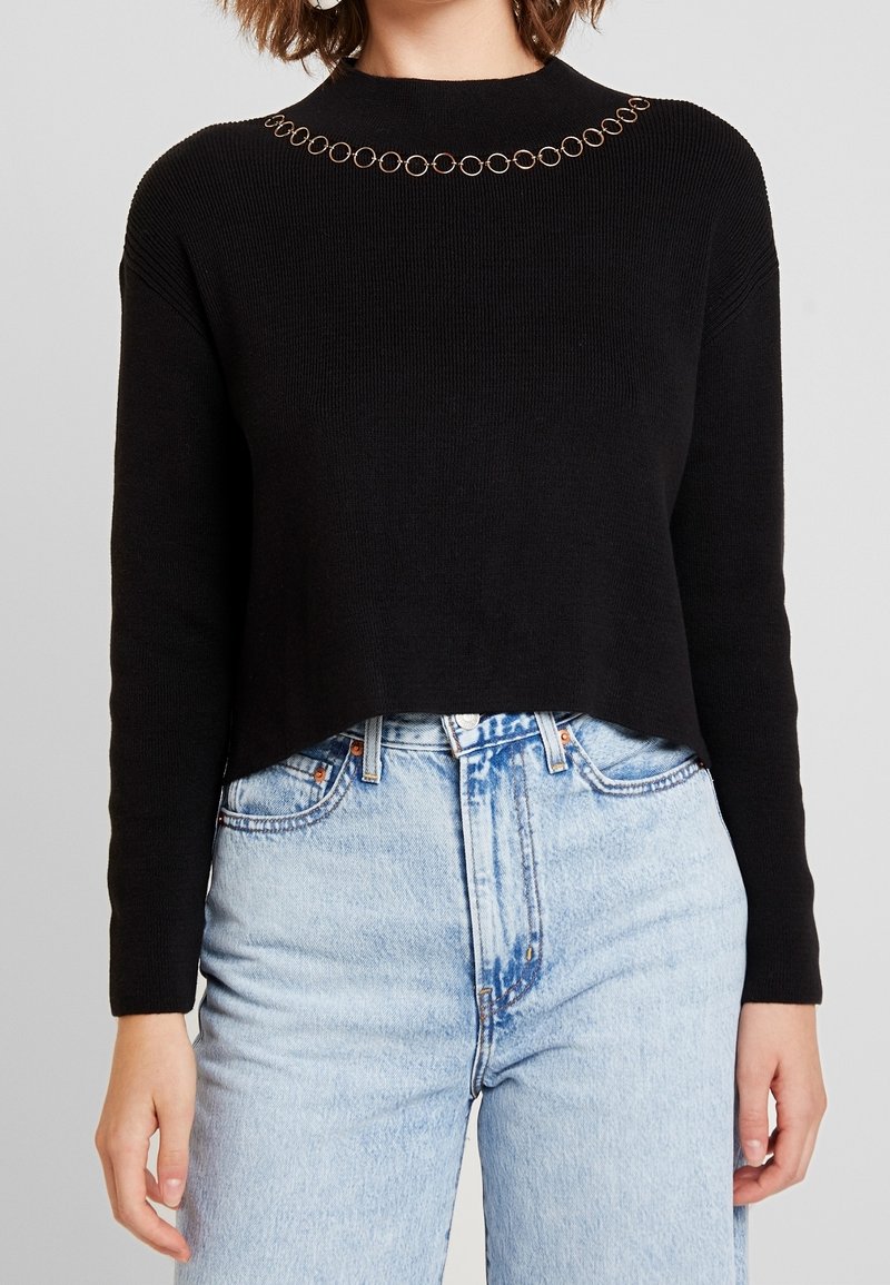 Femme portant un pull court noir à manches longues avec un détail de chaîne circulaire près de l'encolure et un jean bleu clair taille haute.