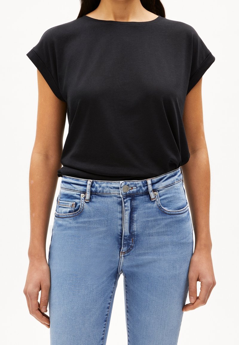 Femme portant un t-shirt noir uni à manches courtes, rentré dans un jean denim bleu clair taille haute, sur fond blanc.