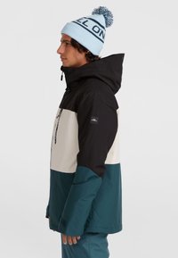 Kleurafgebakende waterdichte jas met een zwart, wit en teal ontwerp; voorzien van een capuchon en rits; aangevuld met een blauwe gebreide beanie met pompon.