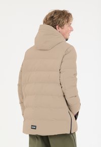 Beige puffjacka med huva, quiltad design, elastiska manschetter och dragkedjefickor. Har en logotagg på baksidan och en slät yta.