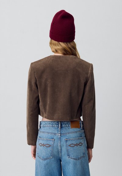 Giacca in velluto corduroy marrone tagliata, con maniche lunghe e scollatura rotonda. Abbinata a jeans blu a vita alta con cuciture decorative sul retro.