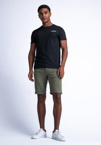 Camiseta de algodón negra con un pequeño logo dorado, combinada con pantalones cortos enrollados de color verde oliva y zapatillas blancas. Diseño simple, ajuste casual.