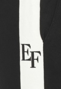 Pantaloni da pista bianco e nero con una striscia laterale, realizzati in un tessuto morbido. Presentano un logo 'EF' nero ben visibile sul lato sinistro.