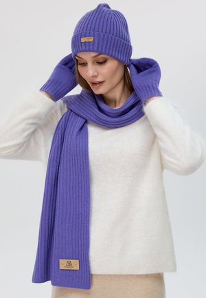 Set di cappello a coste viola, sciarpa e guanti. Presenta una finitura testurizzata e un'etichetta marrone sulla sciarpa. Indossato con un maglione bianco e soffice.