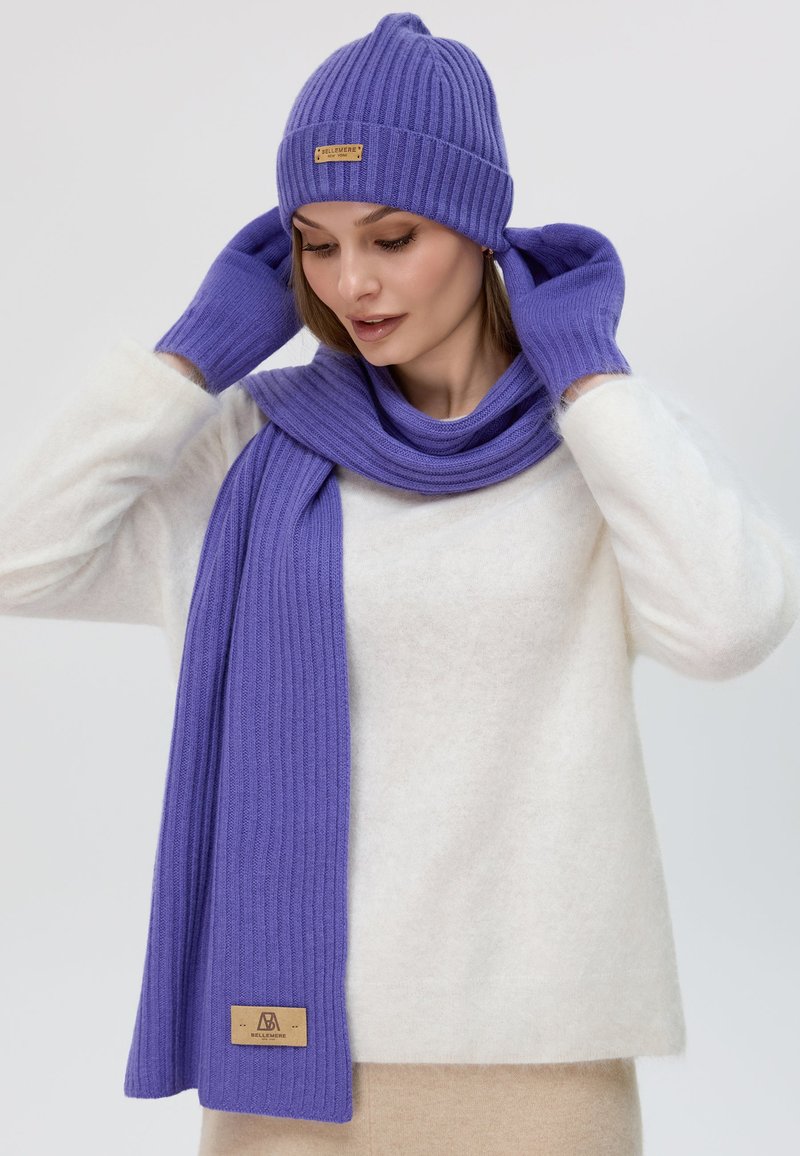 Ensemble comprenant un bonnet, une écharpe et des gants en tricot côtelé violet. Présente une finition texturée et une étiquette marron sur l'écharpe. Porté avec un pull blanc en peluche.