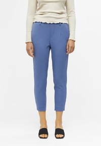 Niebieskie lekko dopasowane spodnie wykonane z elastycznego materiału, z bocznymi kieszeniami i gładkim wykończeniem. Noszone z czarnymi klapkami.