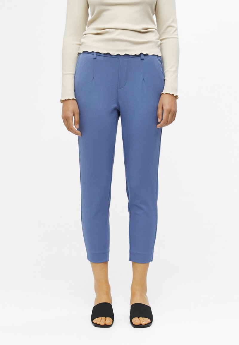 Niebieskie lekko dopasowane spodnie wykonane z elastycznego materiału, z bocznymi kieszeniami i gładkim wykończeniem. Noszone z czarnymi klapkami.