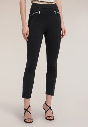 Femme portant un pantalon noir ajusté avec des poches zippées à l'avant et des sandales noires à talons hauts à lanières, debout devant un fond beige.