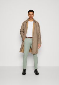 Bruin en beige geblokte overjas, wit t-shirt, lichtgroene broek, zwarte laarzen. Beschikt over twee zijzakken en contrasterende binnenvoering.